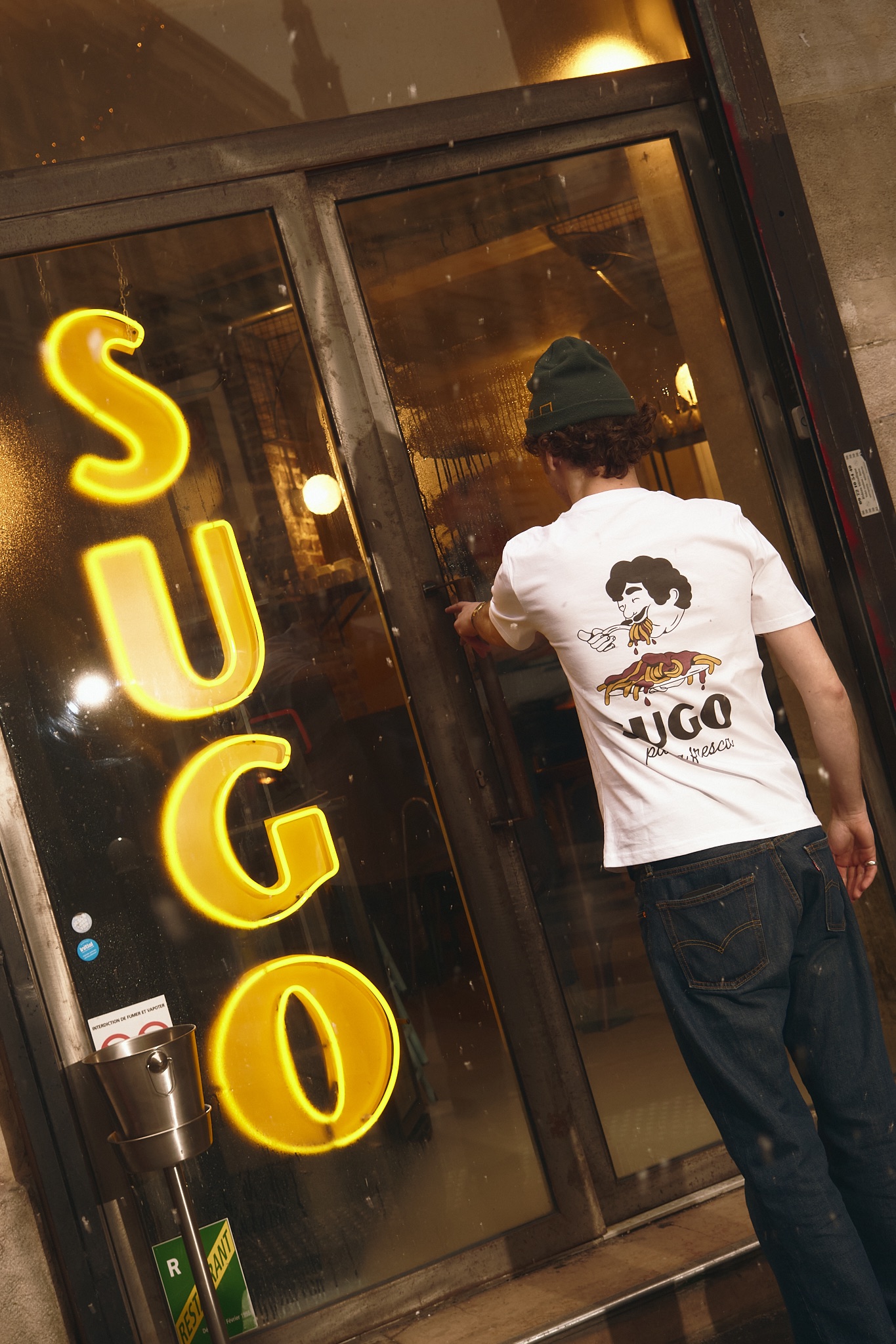 Enseigne néon jaune SUGO sur la vitrine, une personne en t-shirt SUGO pousse la porte